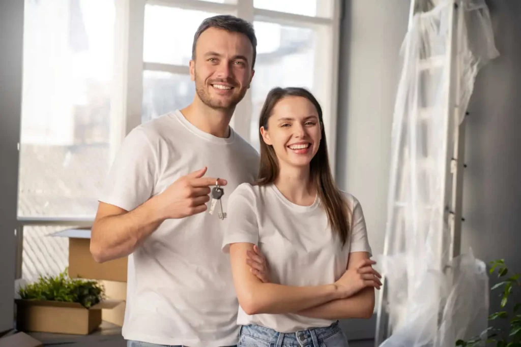 Imagem de um casal sorridente em uma sala iluminada com caixas de mudança e plantas domésticas. O homem segura um molho de chaves e ambos usam camisas brancas casuais, sugerindo que acabaram de se mudar para uma nova casa.