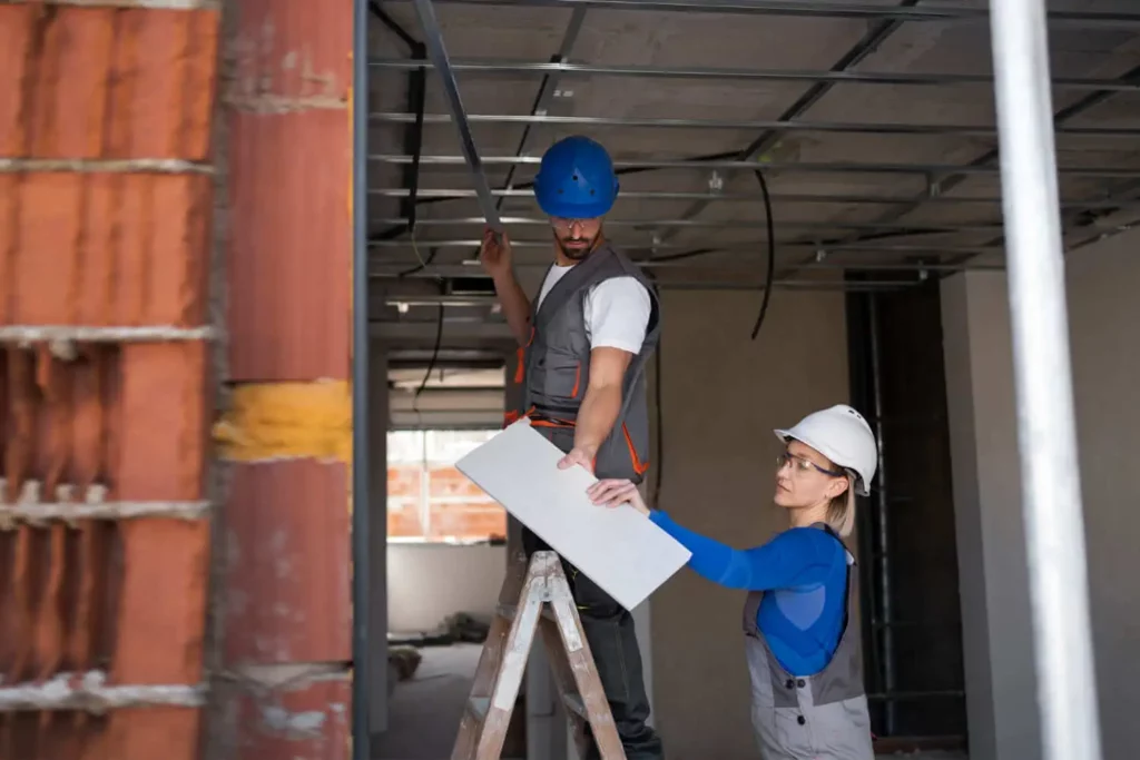 Imagem de dois trabalhadores da construção civil com equipamentos de segurança, um em uma escada segurando um painel de teto e o outro o levantando, trabalham juntos na instalação de painéis em um edifício inacabado. Os tijolos expostos e a estrutura do teto são visíveis.