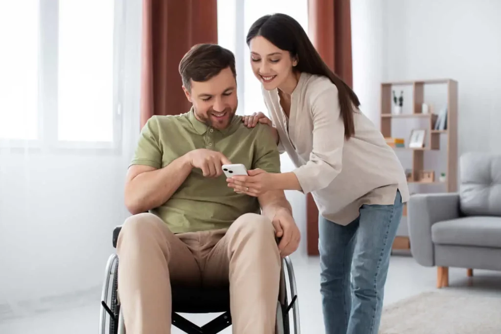 Imagem de um homem em uma cadeira de rodas sorri enquanto segura um smartphone, enquanto uma mulher se inclina sobre seu ombro, também sorrindo e olhando para o telefone com ele em uma sala de estar moderna e iluminada.