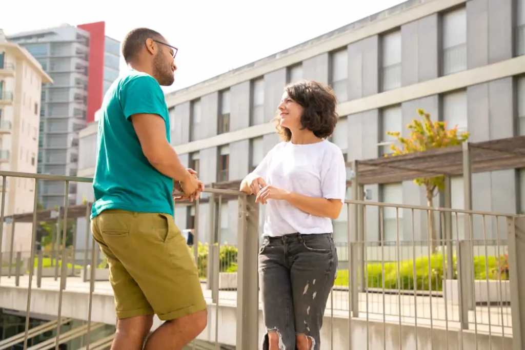 Imagem de duas pessoas ao ar livre em um dia ensolarado, sorrindo e conversando perto de uma grade de metal, com prédios de apartamentos modernos e vegetação ao fundo.