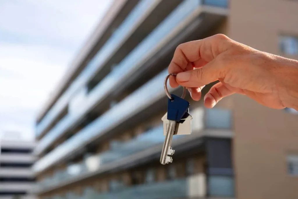 Imagem da mão de uma pessoa segurando a chave de um imóvel em frente a um prédio residencial para ilustrar matéria sobre o que é financiamento imobiliário.