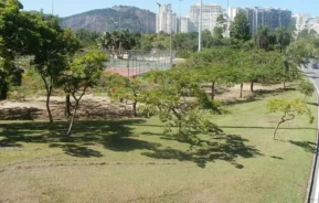 Um parque urbano com grama verde, árvores espalhadas e uma quadra de tênis, tendo como pano de fundo prédios altos e uma colina rochosa sob um céu azul claro.