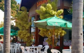 Área de estar externa de uma cafeteria ou restaurante com mesas e cadeiras de plástico branco, guarda-chuvas verdes da Heineken e plantas verdes exuberantes. Um prédio com a placa LARANA está ao fundo em um céu ensolarado.