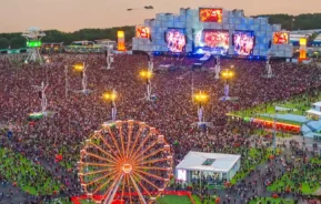 Um grande festival de música ao ar livre ao pôr do sol com uma roda gigante em primeiro plano, uma grande multidão, luzes brilhantes e um palco com telões mostrando os artistas. Árvores e edifícios são visíveis ao fundo.