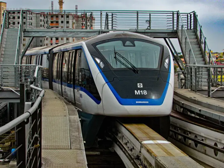 Imagem de um trem de metrô moderno de São Paulo passando por trilhos elevados, cercado por grades de metal e construções urbanas ao fundo.