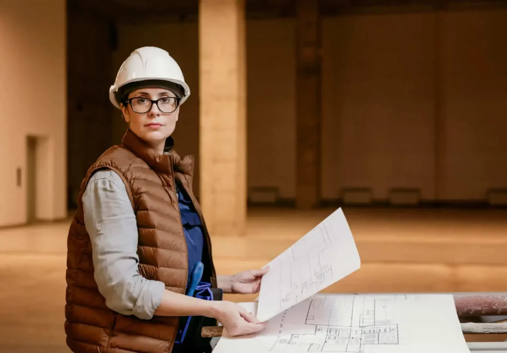 Imagem de uma arquiteta usando capacete, óculos e colete marrom em um local de construção, segurando plantas arquitetônicas e olhando para a câmera para ilustrar matéria sobre o que é RRT de obra. Uma sala grande e vazia e plantas de construção são visíveis.