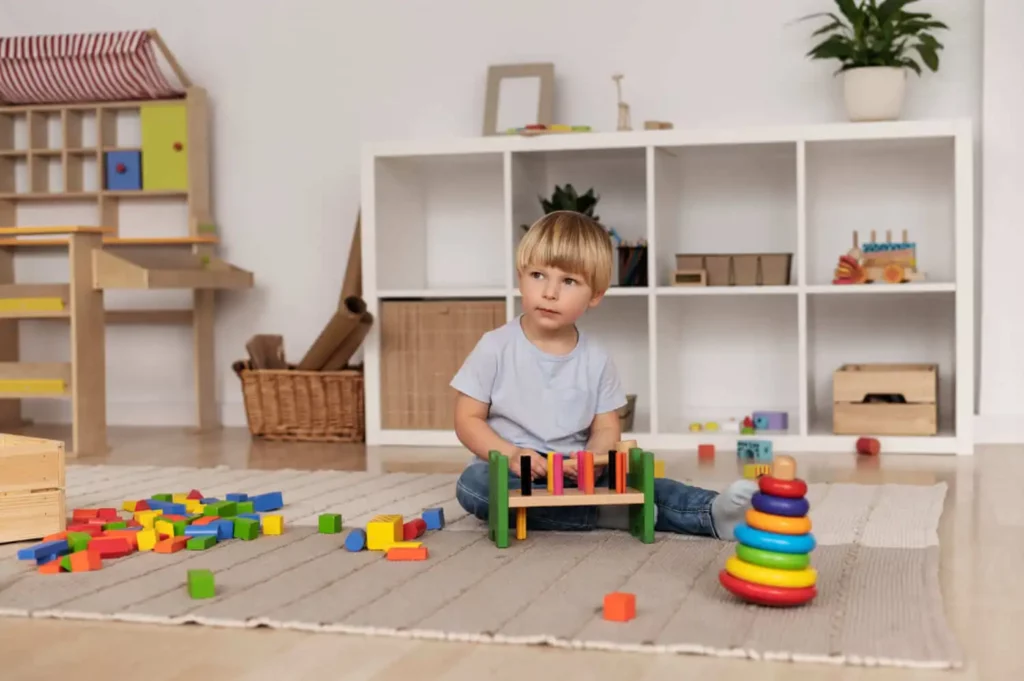 Imagem de uma criança pequena sentada em um tapete em uma sala de jogos, cercada por brinquedos e blocos de madeira coloridos, com prateleiras de cestas e brinquedos ao fundo, para ilustrar matéria sobre o que é uma brinquedoteca