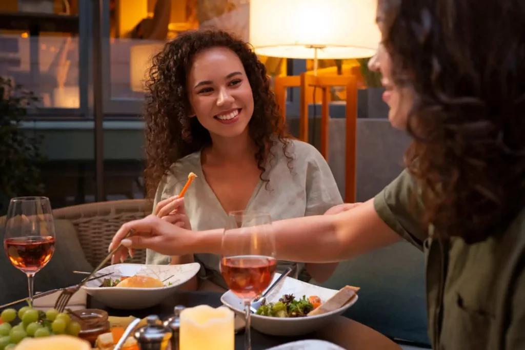 Imagem de duas mulheres sentadas em uma mesa com taças de vinho e pratos de comida, sorrindo e desfrutando de uma refeição juntas, aproveitando o ambiente interno aconchegante e bem iluminado.