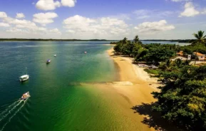 Vista aérea de uma praia tropical com areia dourada, água azul-turquesa clara, barcos flutuando perto da costa e palmeiras ao longo da costa sob um céu azul com nuvens dispersas.