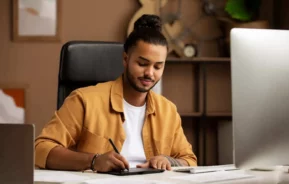 Imagem de um jovem com barba e cabelos presos sentado em uma mesa, sorrindo enquanto usa uma caneta e uma mesa digitalizadora, para ilustrar matéria sobre o Consórcio QuintoAndar. Ele está cercado por papéis, um teclado e um monitor grande em um ambiente de escritório moderno.