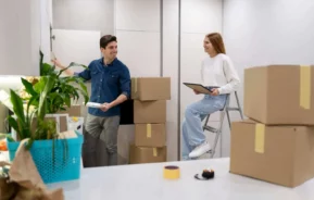 Imagem de um homem e uma mulher desempacotando caixas em um imóvel bem iluminado para ilustrar matéria sobre imóvel comercial. O homem está ao lado de caixas empilhadas, sorrindo e segurando um prato, enquanto a mulher está sentada em uma escada segurando um laptop. Há plantas e caixas de mudança ao redor deles.