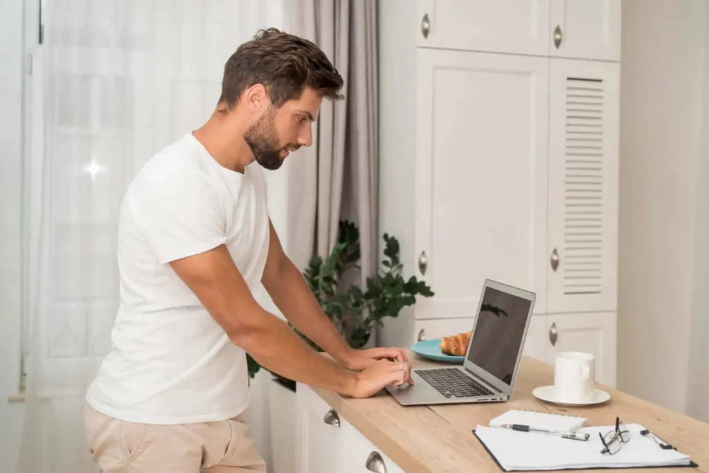Imagem de umhomem de camiseta branca no balcão de uma cozinha usando um laptop para ilustrar matéria sobre o que é subsíndico. Um croissant, uma prancheta e uma xícara de café estão no balcão ao lado dele. Armários brancos e uma planta estão ao fundo.