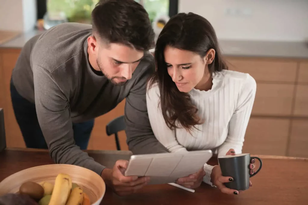 Imagem de um homem e uma mulher em uma cozinha, olhando juntos para um tablet. A mulher segura uma caneca e uma tigela de frutas está sobre o balcão em frente a eles. Eles parecem concentrados na tela.