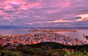 Vista panorâmica de uma cidade litorânea ao pôr do sol, com prédios altos, luzes brilhantes da cidade, vegetação exuberante em primeiro plano e nuvens roxas e rosas dramáticas no céu acima da água e das montanhas.