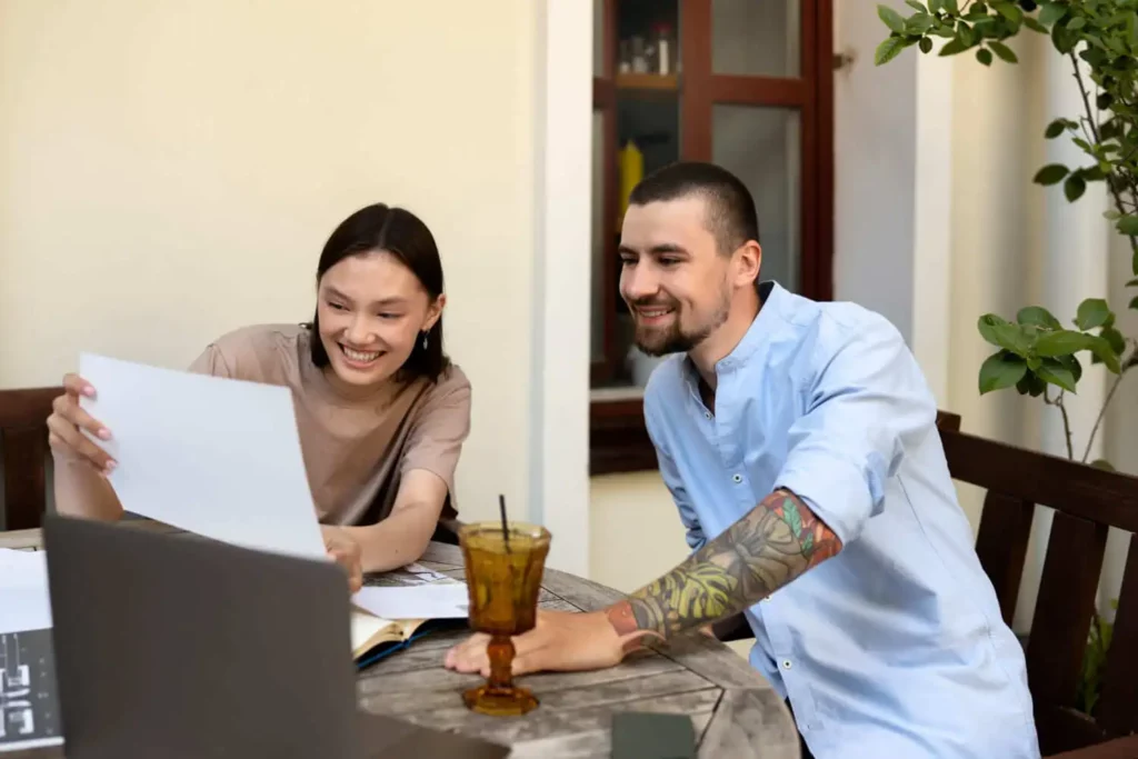 Imagem de duas pessoas sentadas em uma mesa ao ar livre, sorrindo e olhando para documentos em frente a um laptop. Uma bebida em um copo marrom e cadernos de anotações estão sobre a mesa. Plantas e uma janela estão ao fundo.