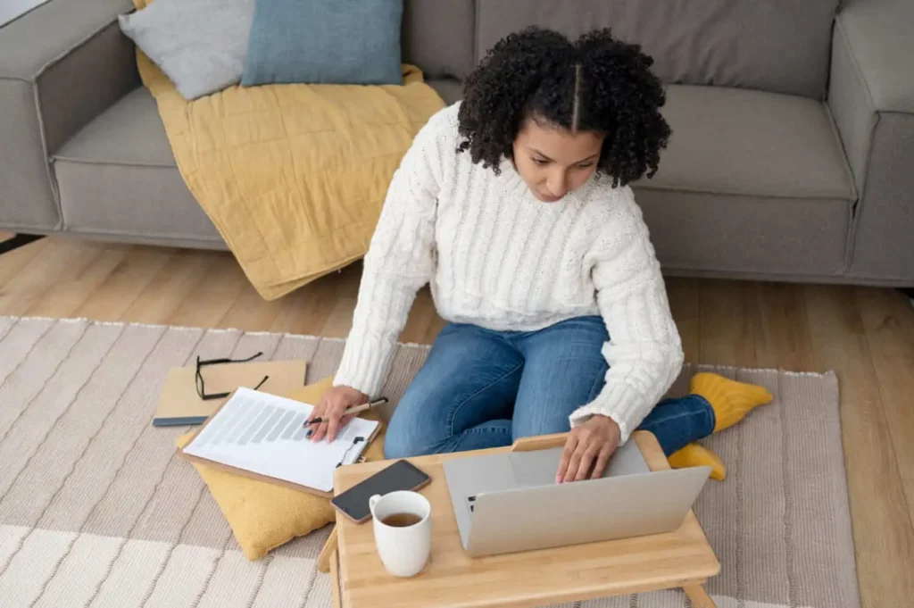Imagem de uma mulher com um suéter branco sentada no chão, trabalhando em um laptop colocado em uma pequena mesa, para ilustrar matéria sobre como reduzir inadimplência no consórcio. Há papéis, uma prancheta, um telefone, óculos e uma caneca de café por perto. Um sofá e um cobertor estão ao fundo.