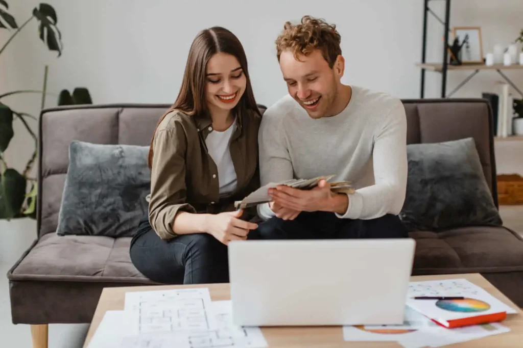 Imagem de um casal sorridente sentado em um sofá, olhando para papéis e um tablet juntos, com um laptop e documentos espalhados em uma mesa de madeira na frente deles em uma sala de estar aconchegante para ilustrar matéria sobre as consequências da inadimplência no consórcio.