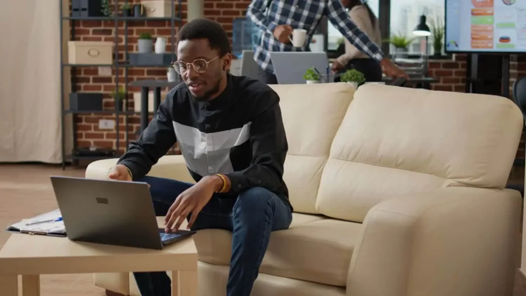 Imagem de um homem de óculos e camisa preta sentado em um sofá bege usando um laptop para ilustrar matéria que explica o que acontece se não pagar o consórcio. Um caderno aberto e uma caneta estão sobre a mesa de centro. Ao fundo, outra pessoa está em uma mesa com computadores em um escritório moderno, com paredes de tijolos.
