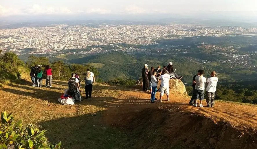 Imagem de um grupo de pessoas no Pico do Urubu, o mirante mais famoso de Mogi das Cruzes. O cenário da cidade é visível ao fundo.