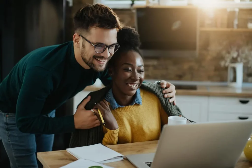 Imagem de um homem sorridente ao lado de uma mulher que está sentada à mesa, ambos olhando para um laptop. A mulher segura uma caneca e há cadernos e uma caneta sobre a mesa em uma cozinha moderna e bem iluminada.