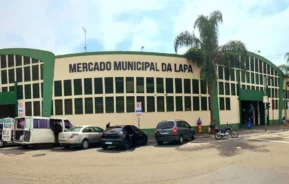 A frente do Mercado Municipal da Lapa, um grande edifício verde e bege com grandes janelas, carros estacionados e pessoas caminhando do lado de fora em um dia ensolarado.