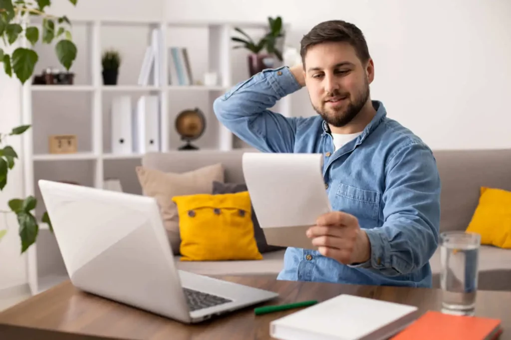 Imagem de um homem sentado em uma mesa em frente a um laptop, segurando um pedaço de papel e coçando a cabeça, parecendo pensativo ou incerto. O plano de fundo mostra um sofá com almofadas amarelas e uma prateleira com decoração.