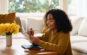 Imagem de uma mulher sorridente com um suéter amarelo sentada em uma mesa de madeira, escrevendo em um tablet com uma caneta stylus, para ilustrar matéria de comparar consórcios. Um vaso de flores amarelas, um telefone e fones de ouvido estão sobre a mesa. Ela parece relaxada em uma sala de estar aconchegante e iluminada pelo sol.