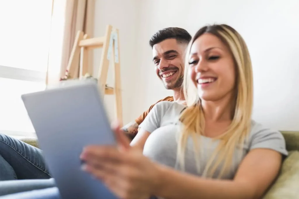Imagem de uma homem e uma mulher sorridentes sentados em um sofá, olhando juntos para um tablet, para ilustrar matéria sobre o que pode e o que não pode para usar o FGTS no consórcio. A mulher segura o tablet em primeiro plano, enquanto o homem está sentado ao lado dela.
