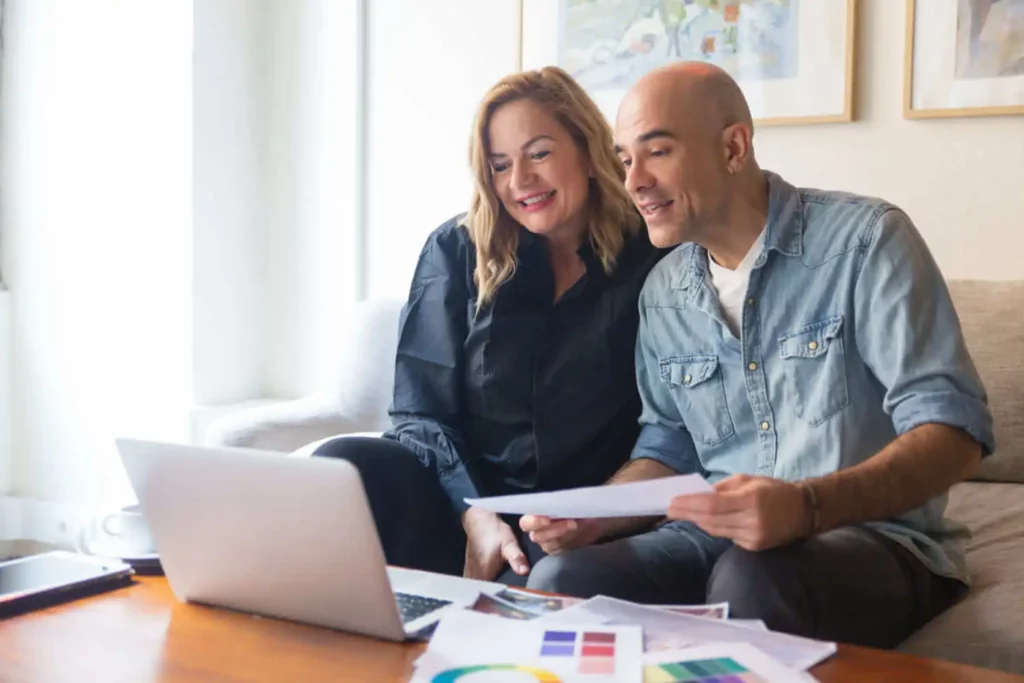 Imagem de um casal sorridente sentado em um sofá, olhando para uma tela de laptop. Eles estão segurando papéis com gráficos coloridos, parecendo engajados e felizes, possivelmente durante uma chamada de vídeo ou uma sessão de planejamento financeiro em casa.