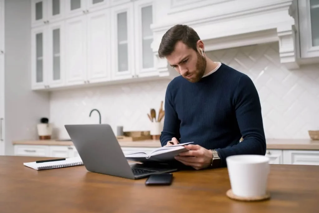 Imagem de um homem vestindo um suéter escuro sentado na mesa da cozinha, escrevendo em um caderno enquanto olha para seu laptop. Um telefone e uma caneca de café branca estão sobre a mesa. A cozinha é moderna e iluminada.