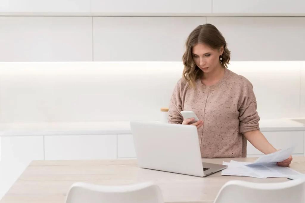 Imagem de uma mulher em uma cozinha moderna, olhando para seu smartphone enquanto segura papéis em uma das mãos e um laptop aberto no balcão à sua frente.