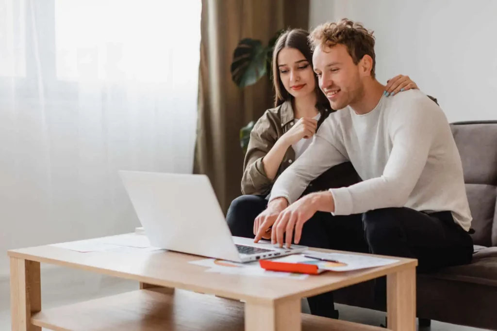 Imagem de um casal sentado junto em um sofá, olhando para um laptop em uma mesa de centro de madeira. O homem sorri enquanto digita, e a mulher pousa gentilmente a mão em seu ombro, ambos parecendo engajados e satisfeitos.
