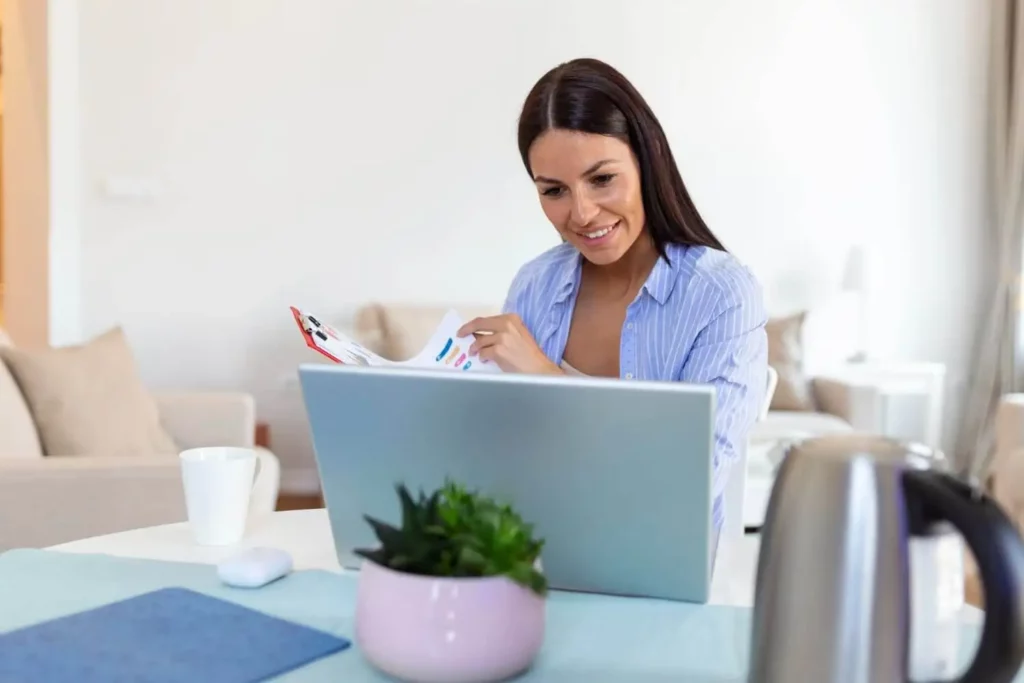 Imagem de uma mulher sentada em uma mesa trabalhando em um laptop, sorrindo enquanto olha para papéis com gráficos coloridos. Uma xícara de café, um vaso de plantas e uma chaleira estão sobre a mesa em uma sala de estar bem iluminada.