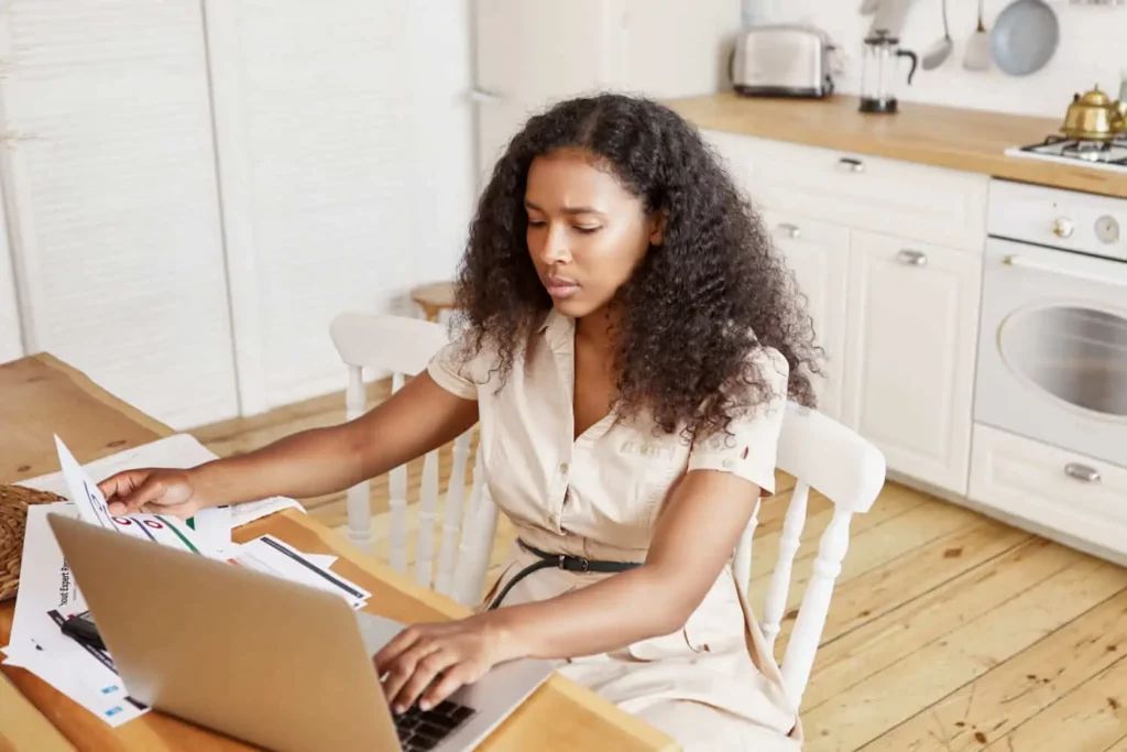 Imagem de uma mulher de cabelos cacheados sentada à mesa da cozinha, trabalhando em um laptop e revisando papéis. A cozinha é bem iluminada, com armários brancos, bancadas de madeira e vários eletrodomésticos ao fundo.