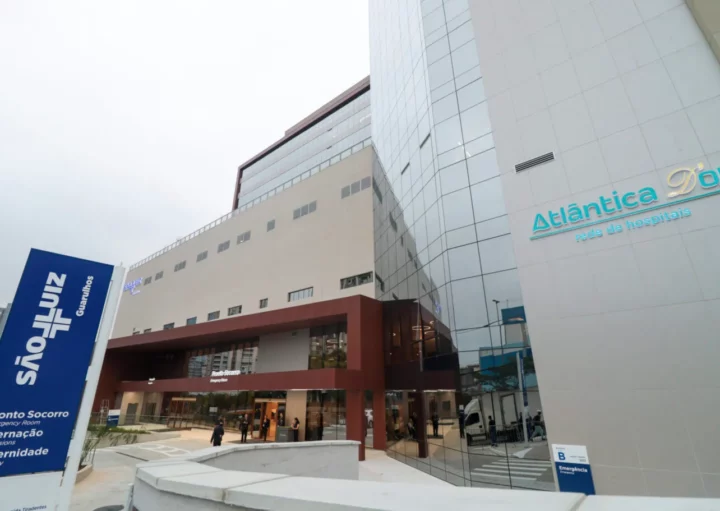 Edifício hospitalar moderno com paredes de vidro e bege. Placas em português mostram Atlântica DOr e Pronto Socorro. As pessoas estão entrando pela entrada principal; a cena se passa durante o dia.