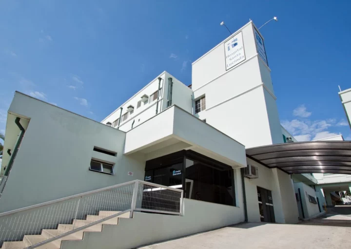 Foto do Hospital Santa Lucinda, em Sorocaba, um edifício moderno, de cor clara, com grandes janelas e uma rampa que leva à entrada, com uma torre com uma placa sob um céu azul claro.