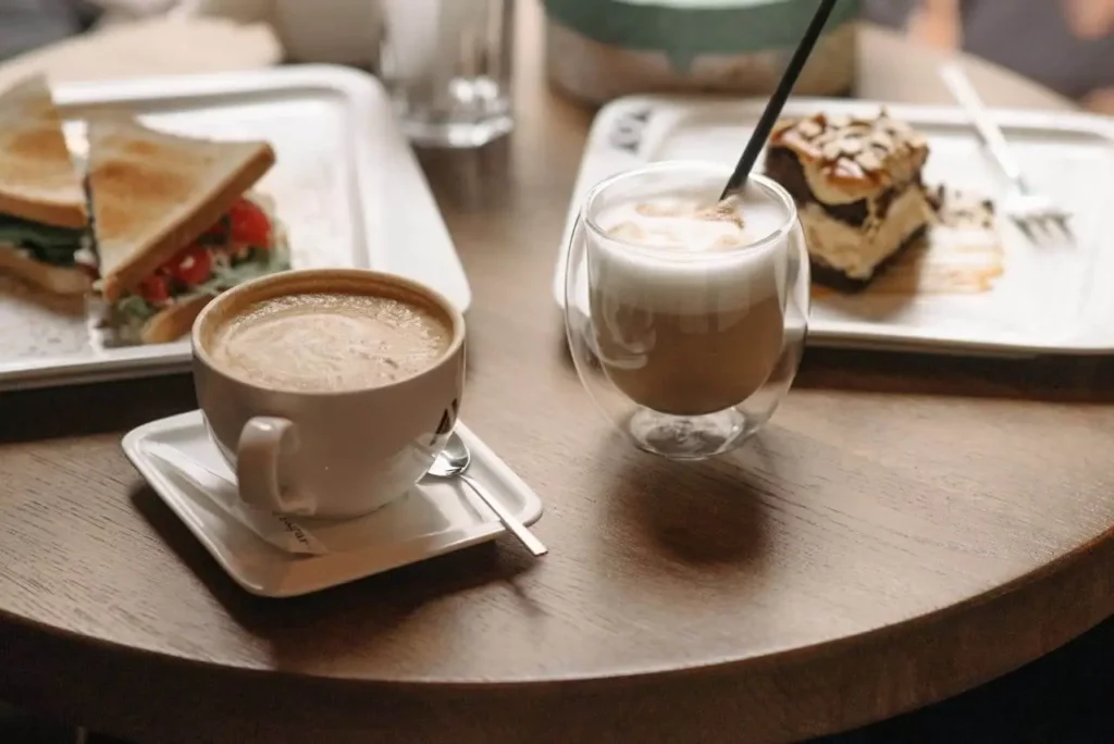 Imagem de uma mesa com uma xícara de café, um copo de cappuccino com canudo, um sanduíche de pão branco com legumes e uma fatia de bolo em camadas coberto com nozes, tudo servido em bandejas, para ilustrar matéria sobre cafeterias boas em Florianópolis.