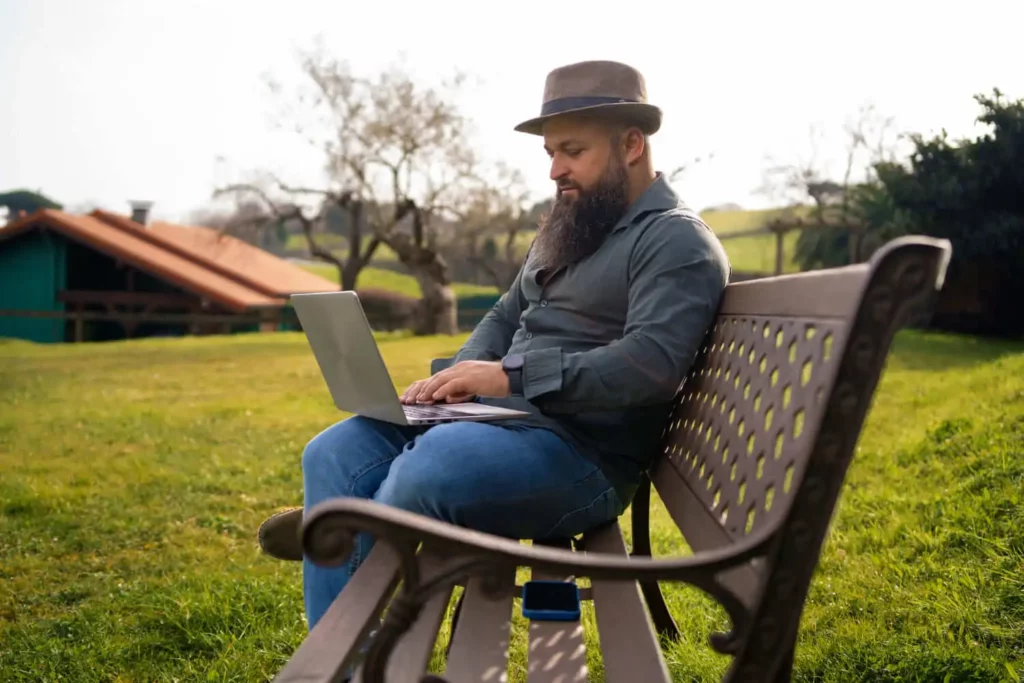Imagem de um homem com barba e chapéu sentado em um banco, trabalhando em um laptop em uma área gramada aberta com árvores e prédios ao fundo.