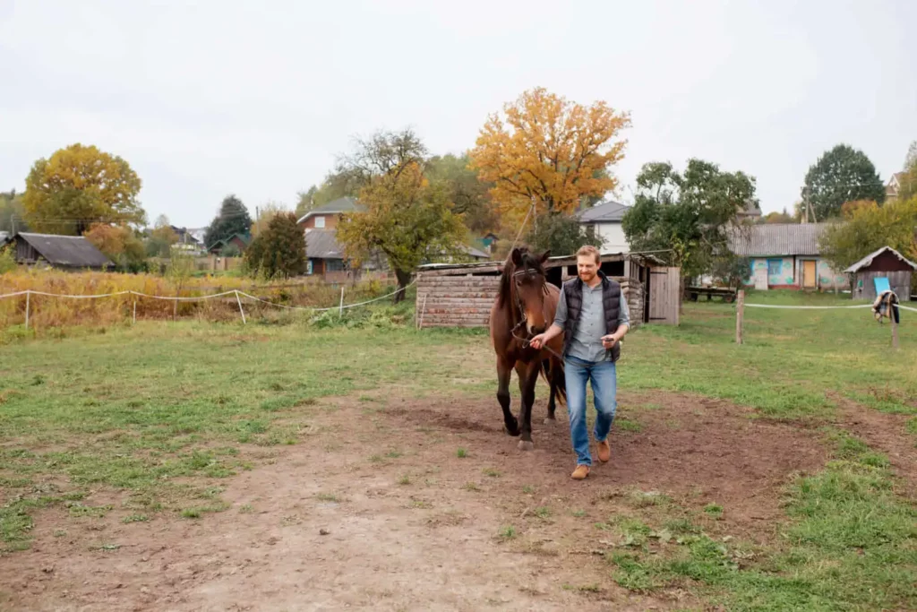 Imagem de um homem de jeans e jaqueta passeando com um cavalo em um pátio gramado com árvores. Ao fundo, há galpões de madeira e casas.