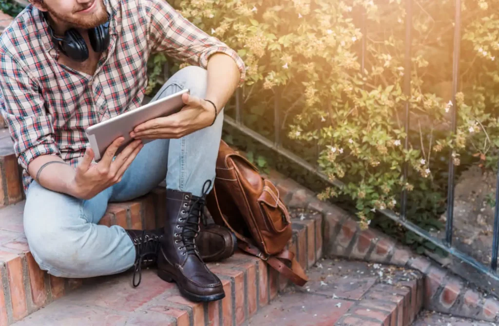 Imagem de uma pessoa de jeans, camisa xadrez e botas sentada em degraus de tijolos ao ar livre, segurando um tablet, para ilustrar matéria sobre CNIR rural. Os fones de ouvido estão em volta do pescoço e uma mochila marrom está ao seu lado. A luz do sol passa por uma vegetação próxima.