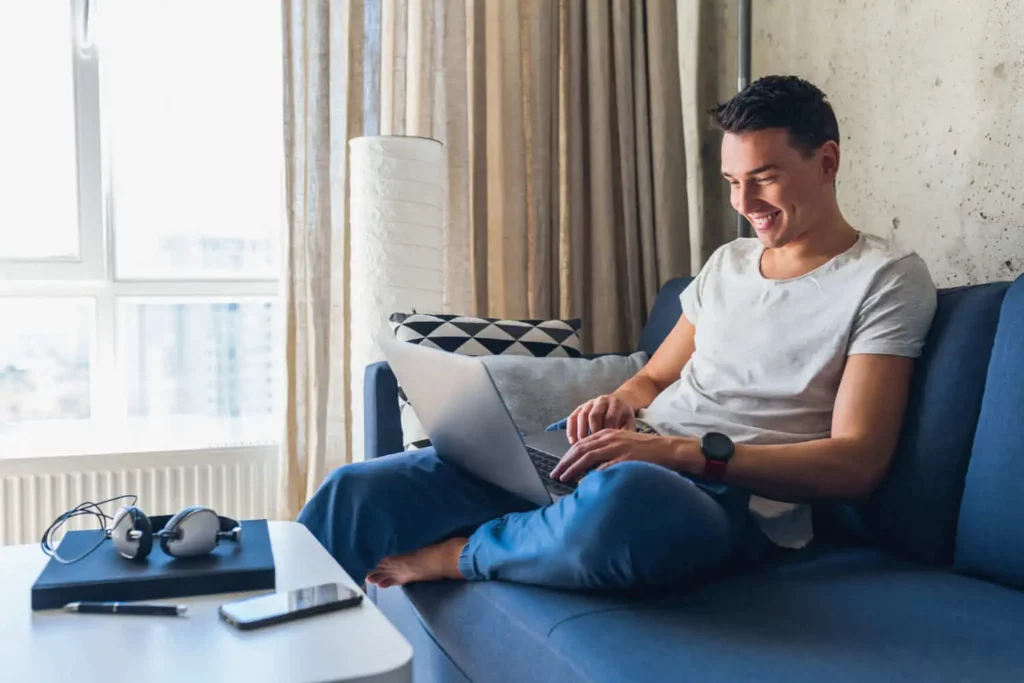Imagem de um jovem sentado em um sofá azul, sorrindo enquanto usa um laptop. Ele está vestido de forma casual e rodeado de aparelhos, incluindo fones de ouvido, um smartphone e um notebook na mesa de centro branca próxima.