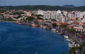 Uma paisagem urbana de um dos bairros de Cabo Frio mostra edifícios e colinas verdes ao fundo, com barcos atracados ao longo de uma estrada costeira ao lado de águas azuis calmas sob um céu claro.