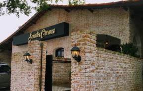 Um prédio rústico de tijolos com um telhado de telhas e uma placa indicando Santa Crema acima da entrada. Luzes em estilo de lanterna estão montadas na parede de tijolos perto de um portão preto. As árvores são visíveis acima da linha do telhado.