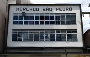 A fachada de um edifício branco com grandes janelas de vidro e o letreiro Mercado São Pedro exibido em letras pretas em negrito no topo.