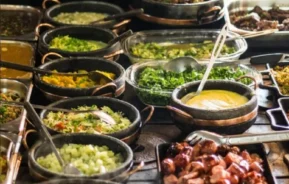 Uma mesa de bufê repleta de vários pratos em potes e bandejas, incluindo legumes, ensopados, arroz, carnes grelhadas, molhos e saladas, todos dispostos próximos uns dos outros e prontos para serem servidos.