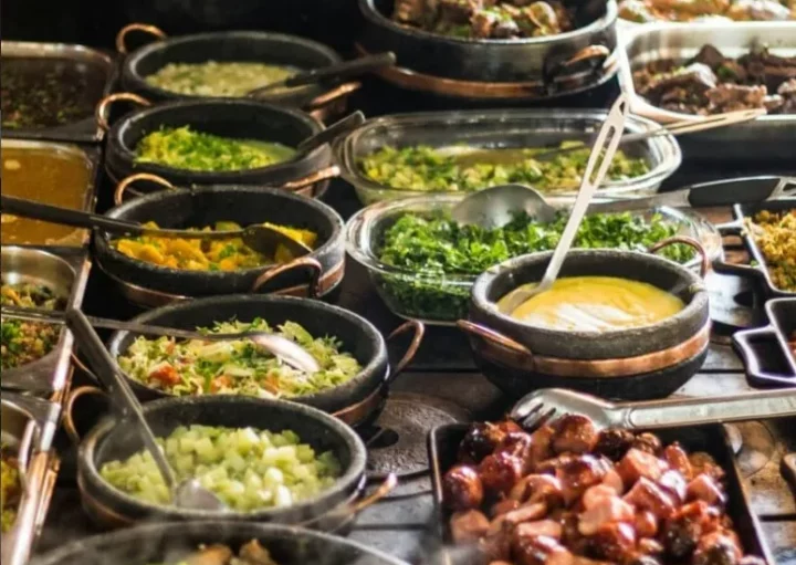 Uma mesa de bufê repleta de vários pratos em potes e bandejas, incluindo legumes, ensopados, arroz, carnes grelhadas, molhos e saladas, todos dispostos próximos uns dos outros e prontos para serem servidos.