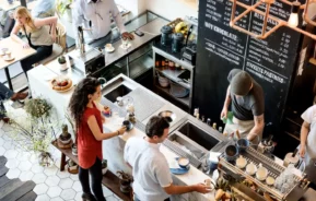 Imagem de uma cafeteria movimentada vista de cima, com baristas preparando bebidas atrás do balcão, clientes esperando e conversando, e pessoas sentadas em mesas próximas a um grande cardápio na parede, para ilustrar matéria sobre cafeteria em Campinas
