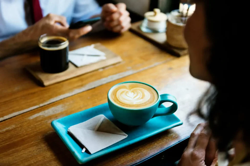 Imagem em formato close-up de uma mesa em um café com uma xícara azul de café com leite e dois guardanapos em uma bandeja, e outra pessoa com uma xícara de café preto ao fundo, ambas sentadas em uma mesa de madeira, para ilustrar matéria sobre cafeteria gourmet em Campinas