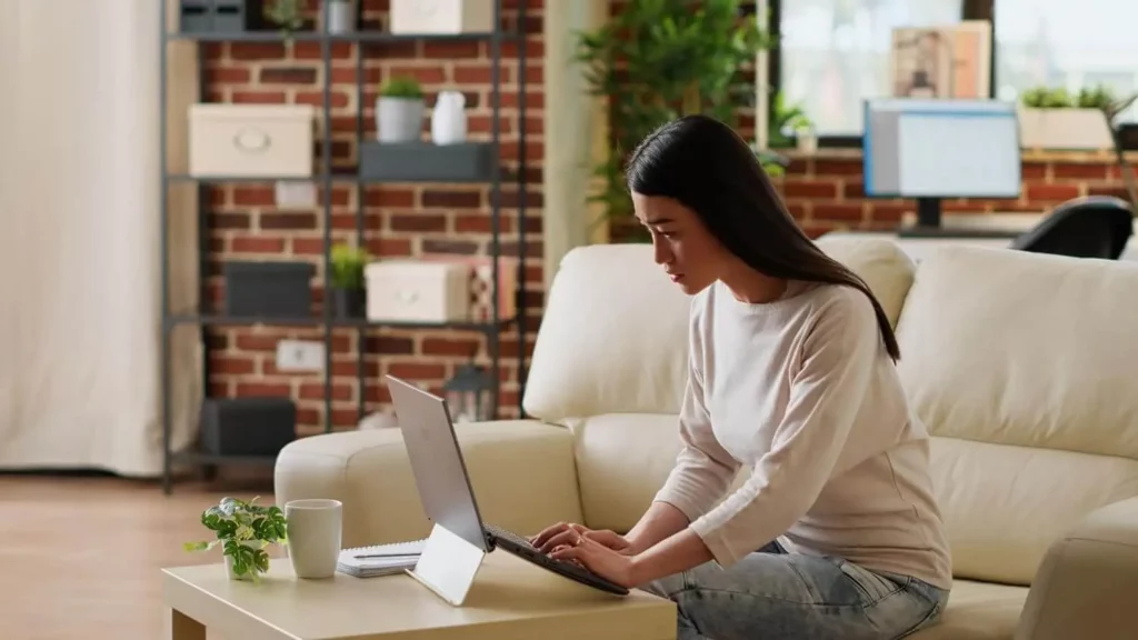 Imagem de uma mulher sentada em um sofá bege em uma sala de estar moderna, digitando em um notebook que está em cima de uma mesa de centro, para ilustrar matéria sobre como desistir do consórcio. Uma xícara e uma pequena planta também estão sobre a mesa, e prateleiras e uma escrivaninha podem ser vistas ao fundo.
