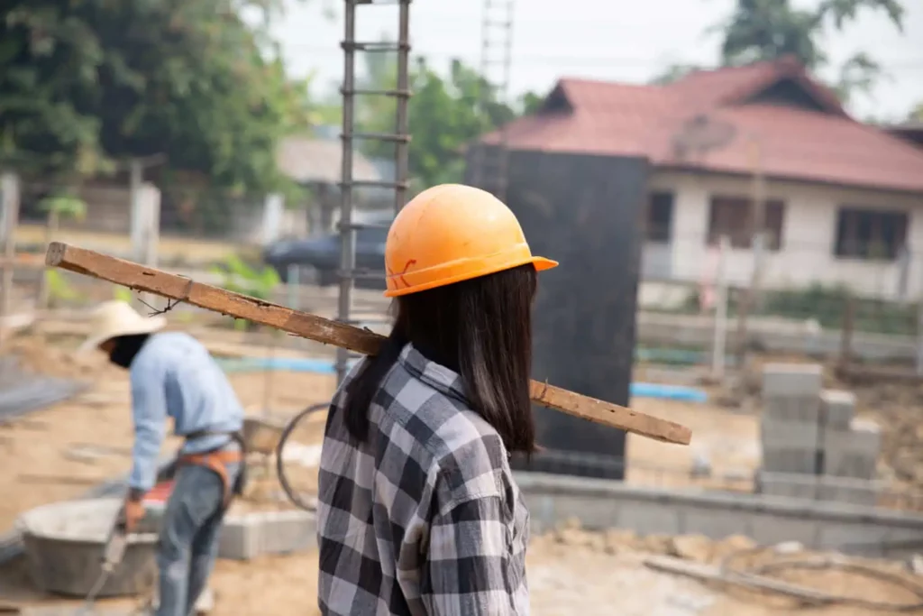 Imagem de um trabalhador da construção civil usando capacete laranja e camisa xadrez carrega uma prancha de madeira no ombro, com outro trabalhador e materiais de construção visíveis ao fundo.